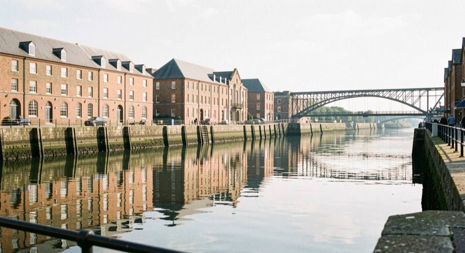 Exeter Quay on a bright morning with the historic warehouses reflected in the river and a footbridge beyond.